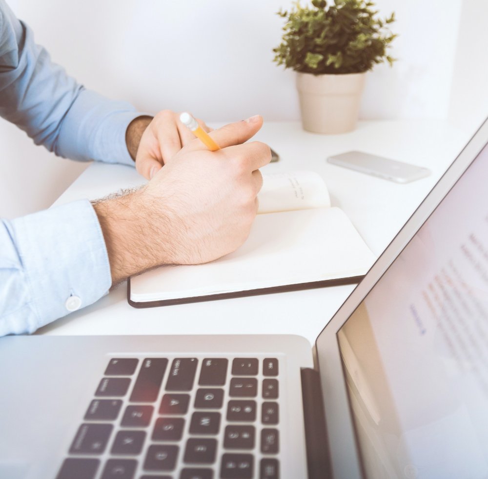 Person holding pencil and writing in notepad, laptop beside them